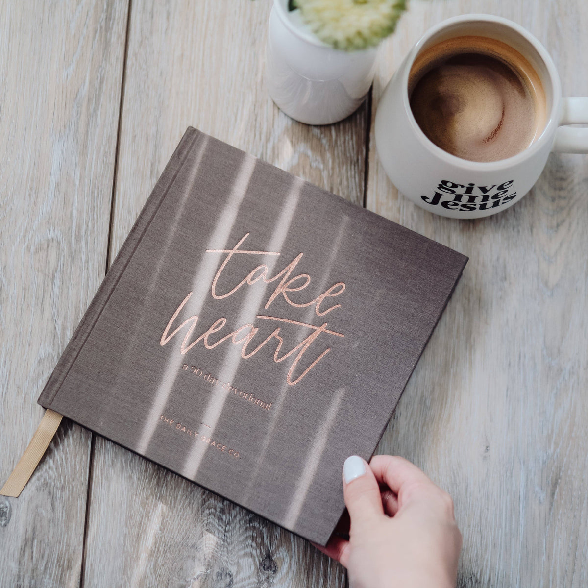 Take Heart devotional book with gray cover and rose gold lettering, positioned on whitewashed wood table with coffee mug and potted plant nearby