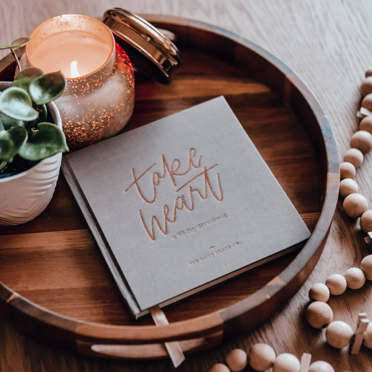 Take Heart devotional book with cream cover and rose-gold lettering on wooden tray, surrounded by sparkly rose-gold candle, white potted succulent, wooden beads, and cinnamon sticks