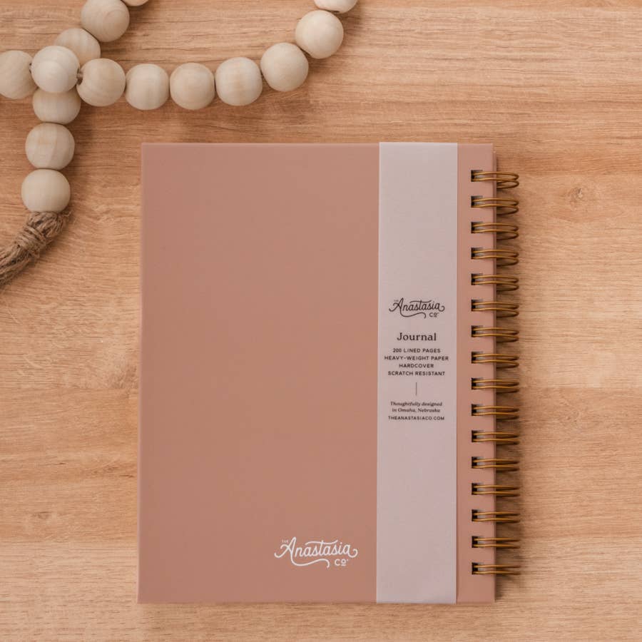 Dusty rose hardcover spiral-bound journal with cream band and brass binding, styled on wood with cream beaded garland