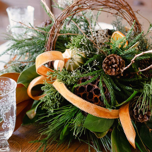 Decorative wreath with greenery, pinecones, and orange ribbon on a table.