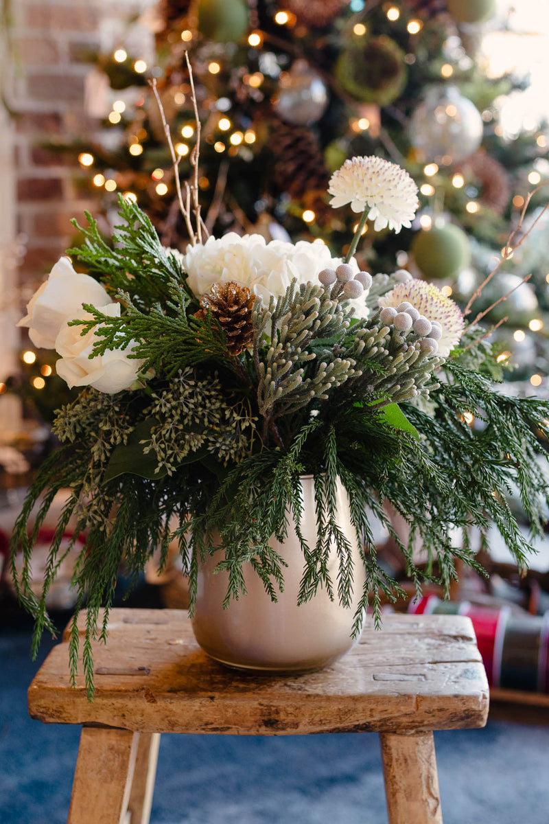Decorative floral arrangement with white flowers and greenery on a wooden stool in front of a Christmas tree.