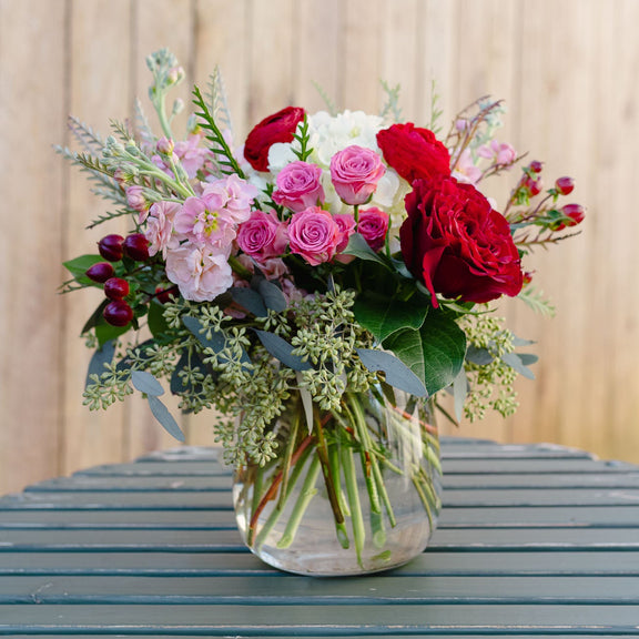 Bouquet of red, pink, and white flowers in a clear vase on a wooden surface.