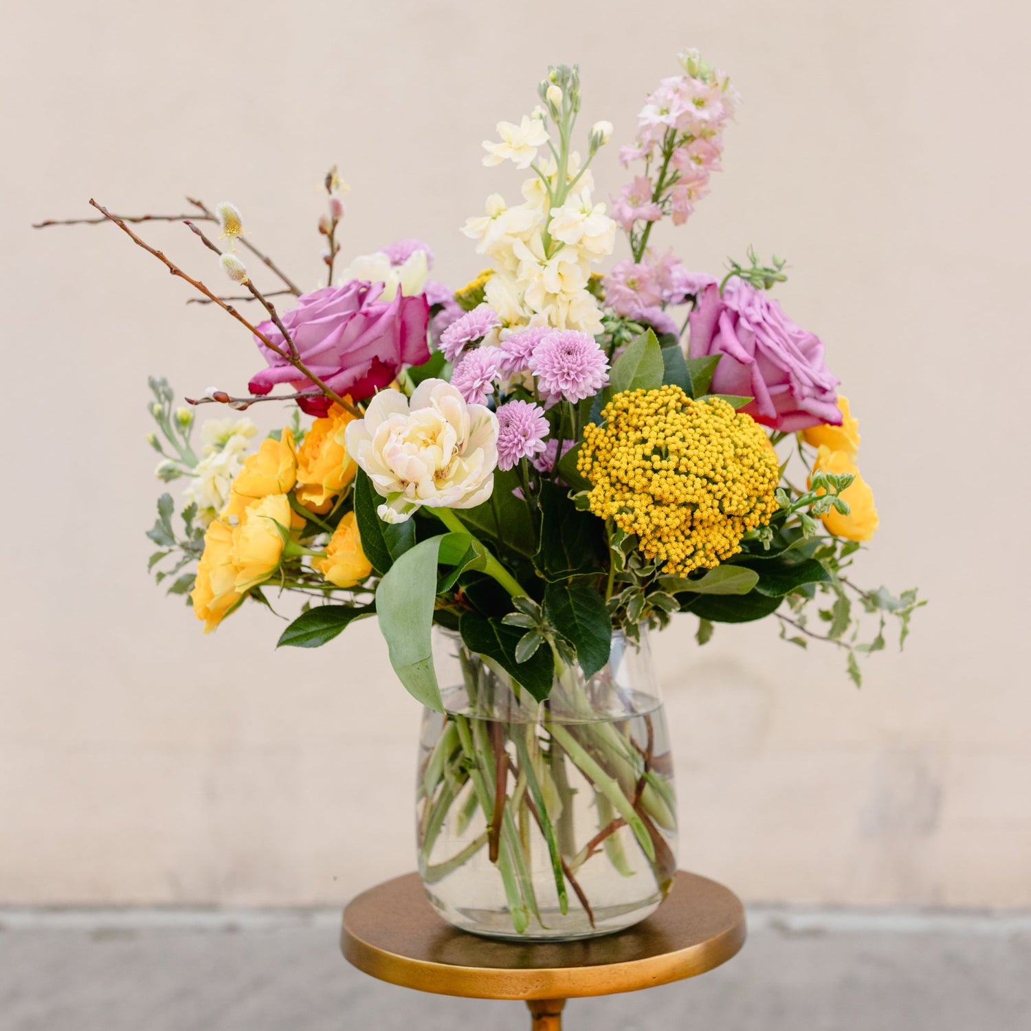 Colorful flower arrangement in a glass vase on a small round wooden table against a plain background