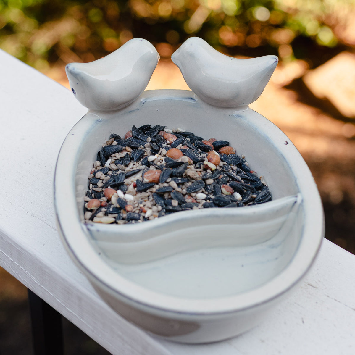 White ceramic bird bath with two curved bird perches and seed basin filled with dark birdseed, positioned in a garden setting