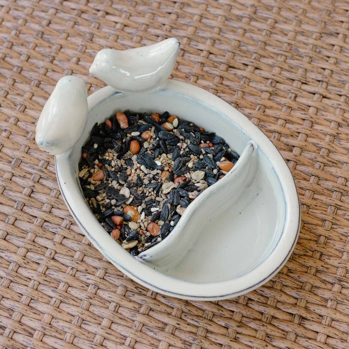 White ceramic bird bath with dual shallow basins and perched ceramic bird finial on top, filled with seed mix.