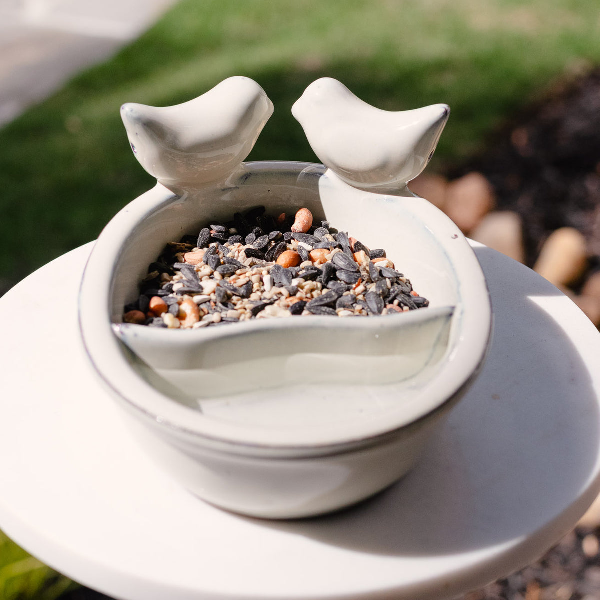 White ceramic bird bath and feeder with two sculptural bird perches, central basin filled with mixed black and tan birdseed, on pedestal in garden