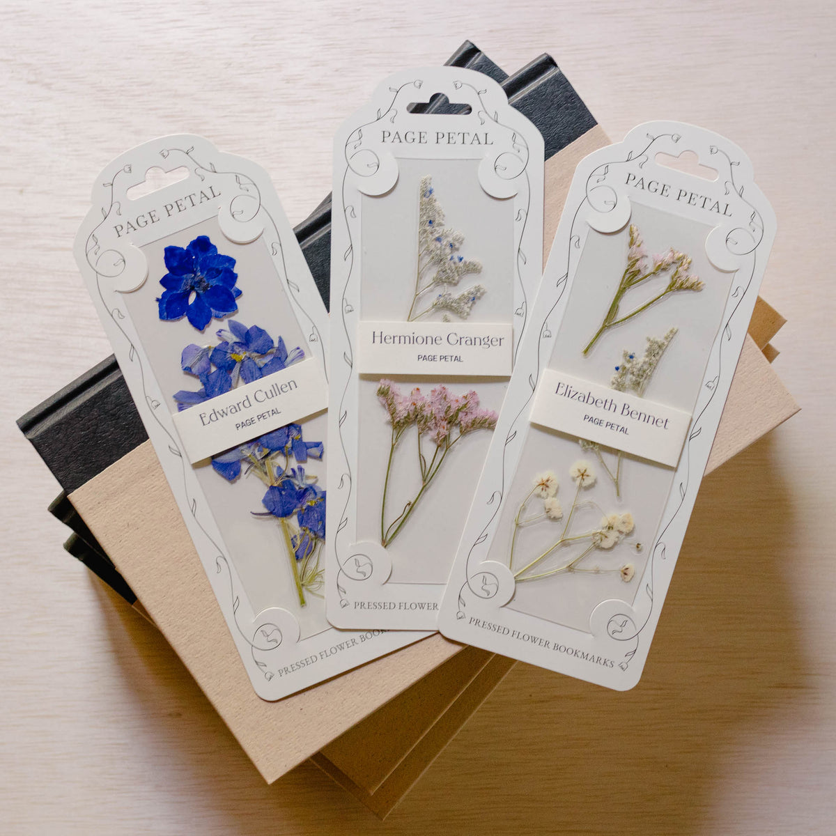 Three Page Petal floral bookmarks fanned out, each displaying pressed flowers and botanical illustrations in blue, white, green, and pink tones