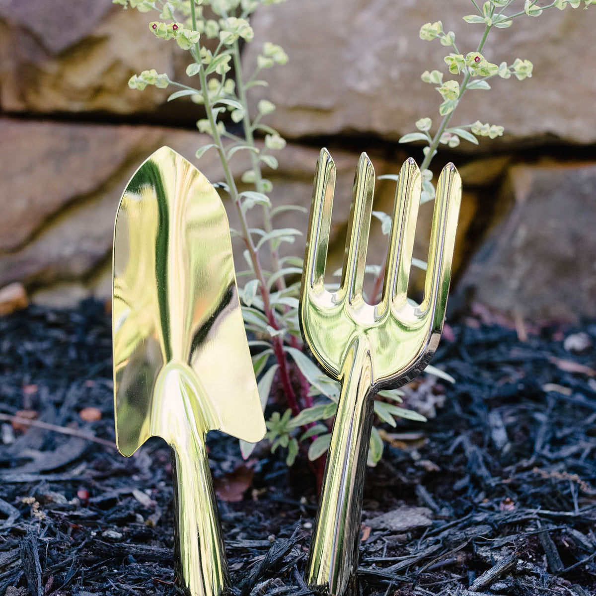 Gold-finish steel garden shovel and hand rake planted in dark soil with delicate white flowers blooming from their handles