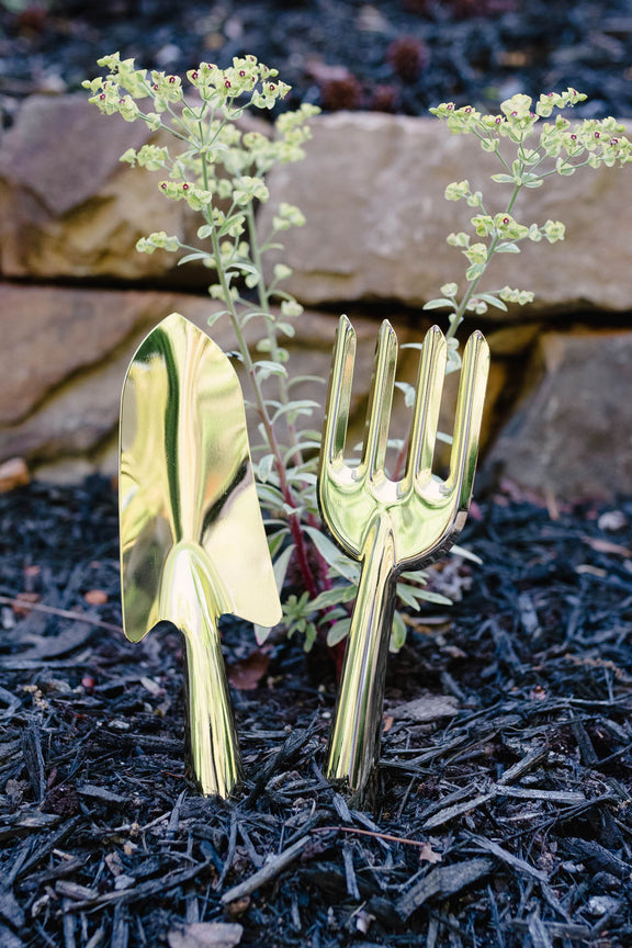 Gold-finish steel garden shovel and hand rake planted in dark soil with delicate white flowers blooming from their handles