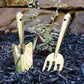 Gold-finish steel shovel and hand rake with long handles standing upright in dark mulch beside a small green plant.