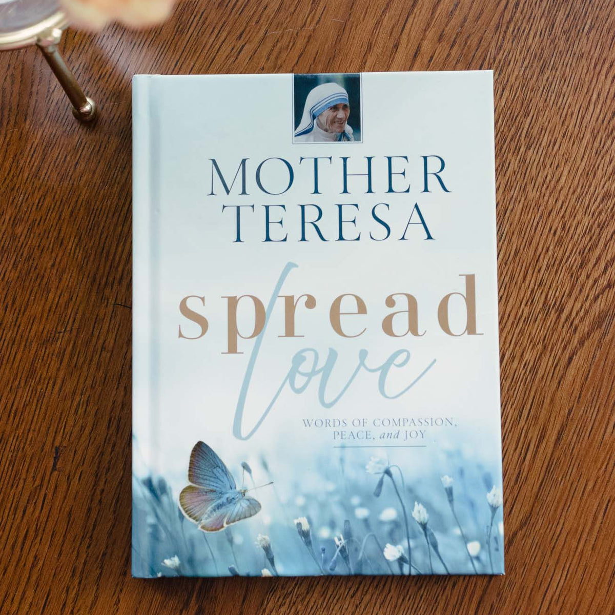 Hardcover book 'Mother Teresa: Spread Love' with blue watercolor flowers and butterfly on cover, displayed on wood with peach dahlia.