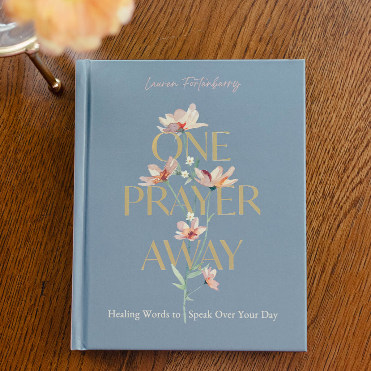 Blue hardcover book "One Prayer Away" with gold lettering and pink floral design, next to a peach dahlia on wood surface.