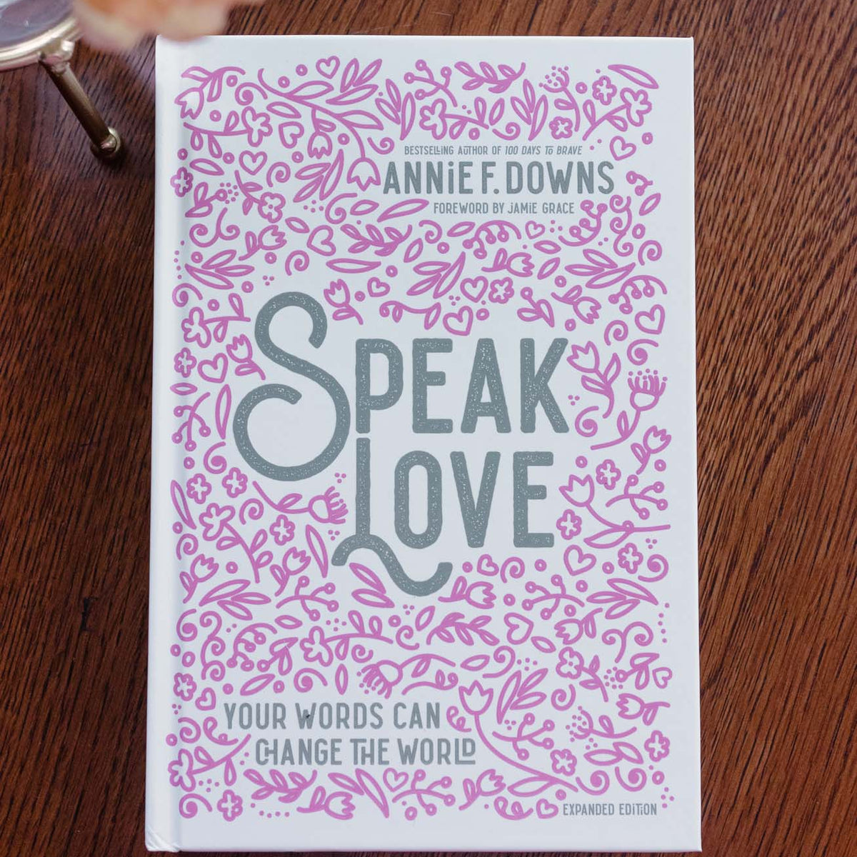 "Speak Love" by Annie F. Downs book cover with pink ornamental pattern on wood surface, peach flower nearby
