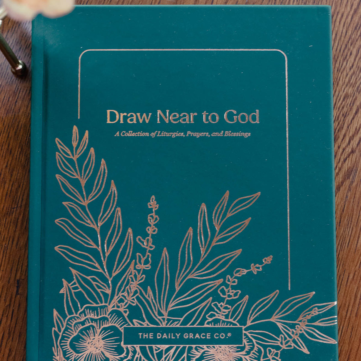 Teal blue hardcover book titled "Draw Near to God" with copper botanical line art illustrations and pale dried flower on wood surface.