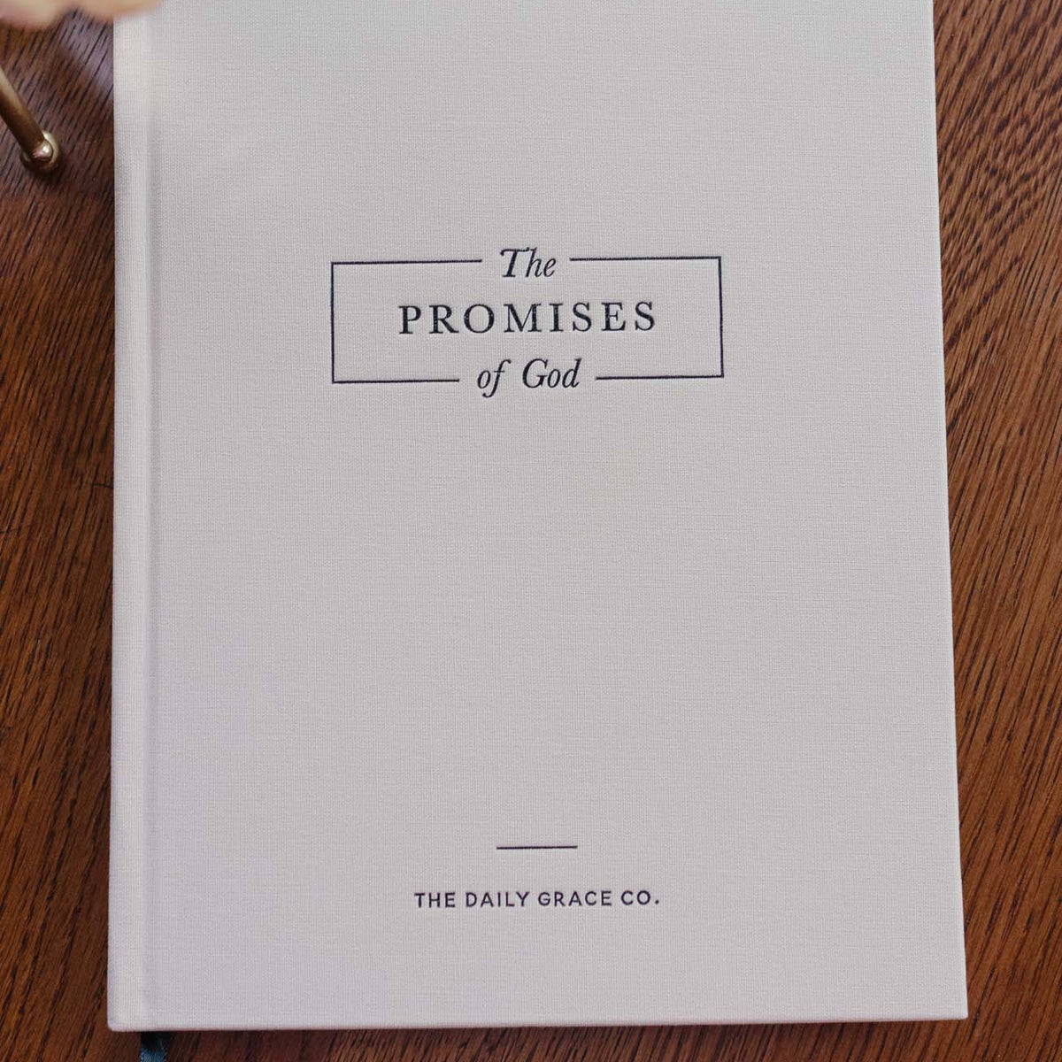 Cream hardcover book titled 'The Promises of God' by The Daily Grace Co on wooden surface with peach flower and blue ribbon bookmark