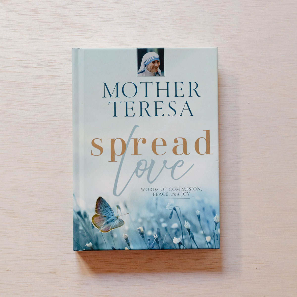 Hardcover book with pale blue cover featuring Mother Teresa's portrait, title in teal, "spread love" in copper and blue script, and blue butterfly on wildflowers at base.