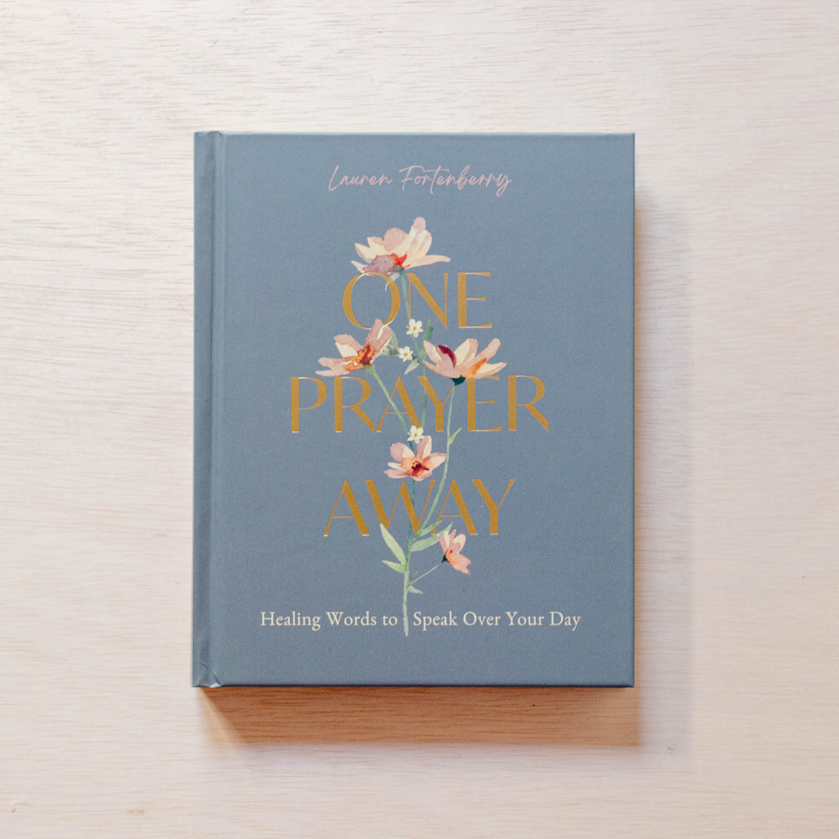 Blue hardcover book 'One Prayer Away' by Lauren Fortenberry with watercolor flowers and gold lettering on cover