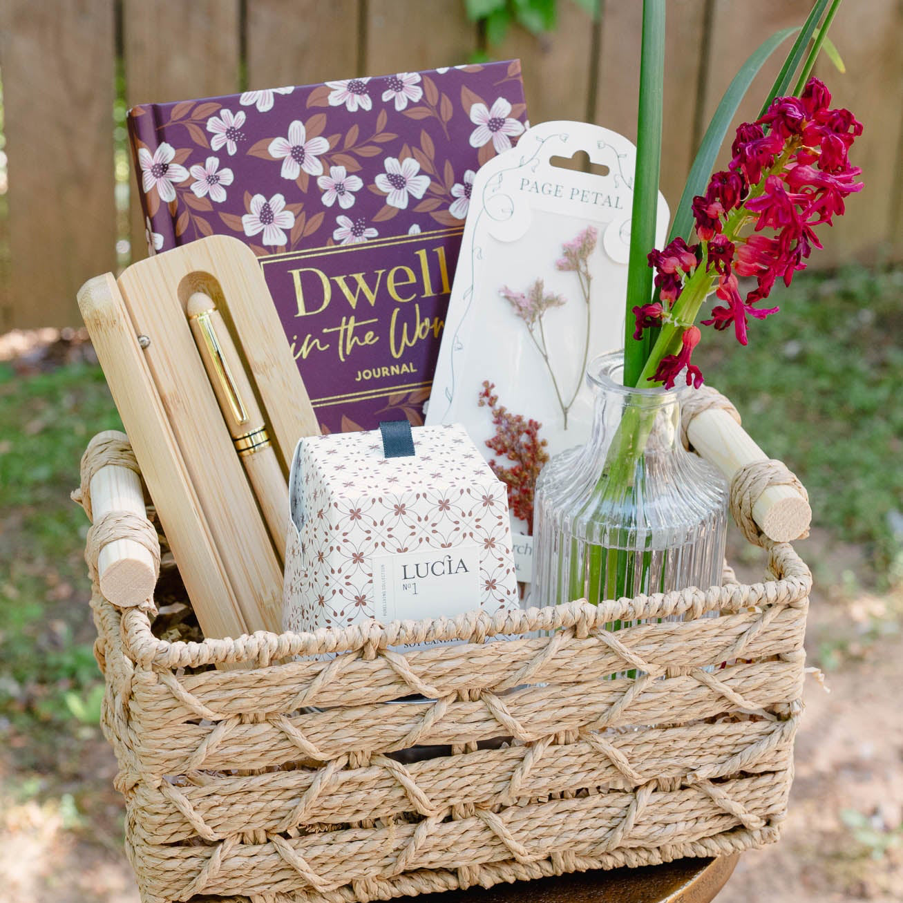 Woven basket with decorative items including a book, a small plant, and a candle on a wooden stand outdoors.