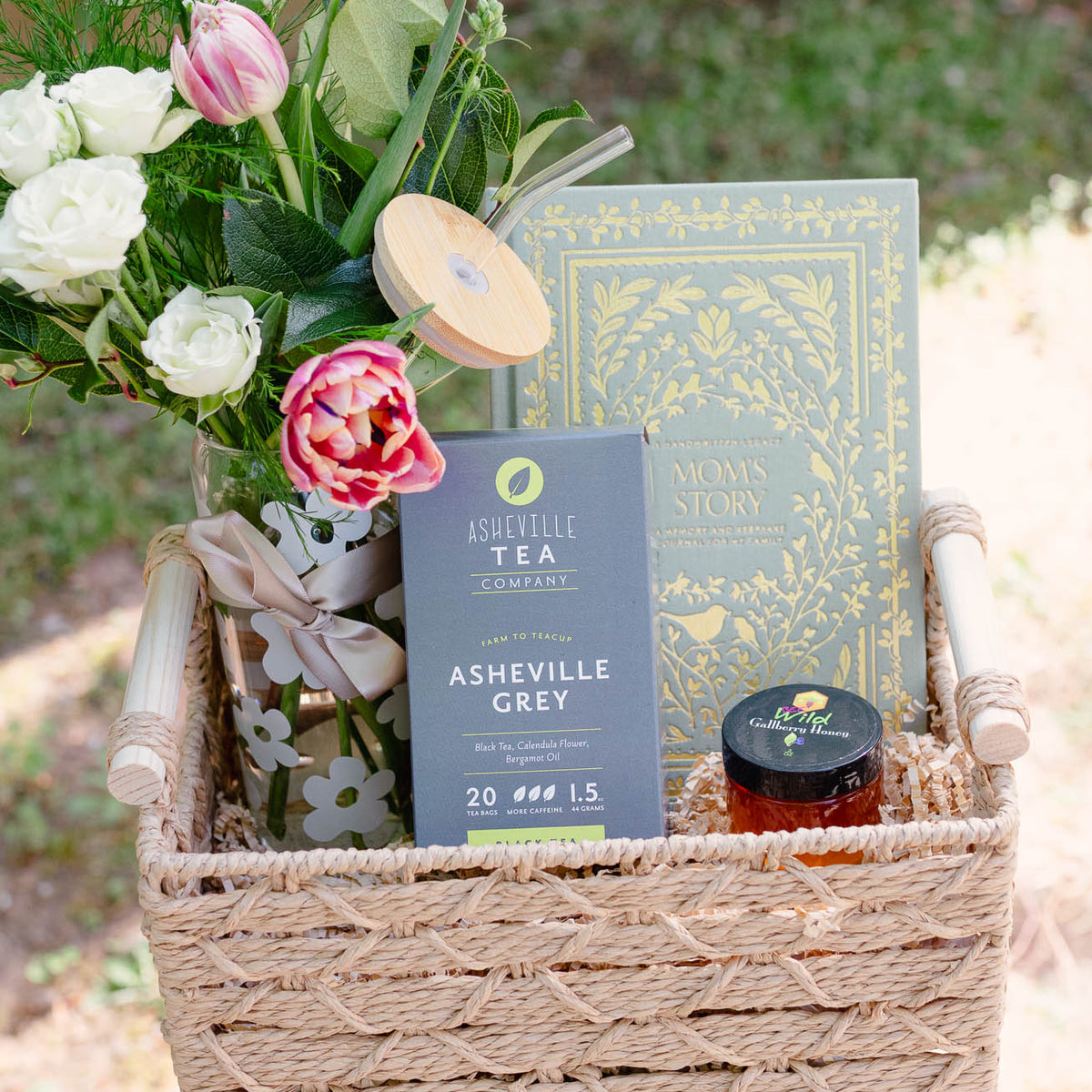 Woven basket gift set with fresh white roses and pink peonies, navy and gold-embellished journals, amber glass tea jar, and wooden serving paddle