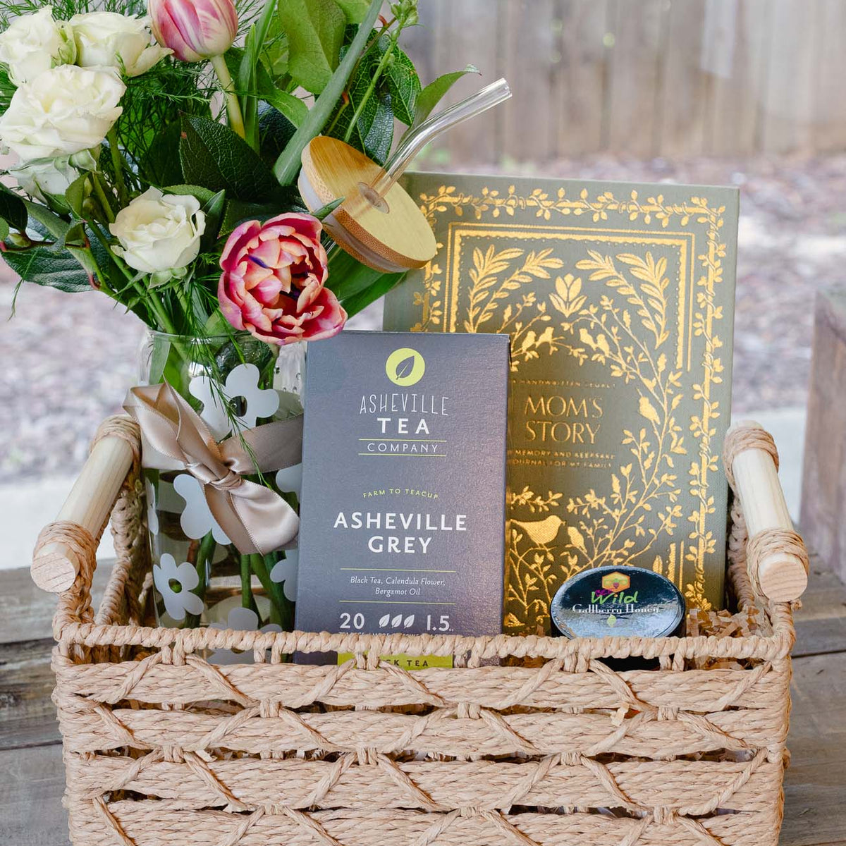 Woven straw basket filled with Asheville Grey tea, Mom's Story journal, and fresh white roses with pink peonies and gold ribbon