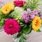 Vibrant floral arrangement with magenta roses, yellow gerbera daisies, pink sweet peas, and green ferns in a clear glass vase