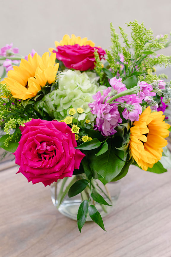 Vibrant floral arrangement with magenta roses, yellow gerbera daisies, pink sweet peas, and green ferns in a clear glass vase