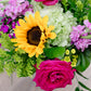 Bright floral arrangement with golden sunflower, magenta roses, pink phlox, green hypericum berries, and cascading greenery on weathered wood surface