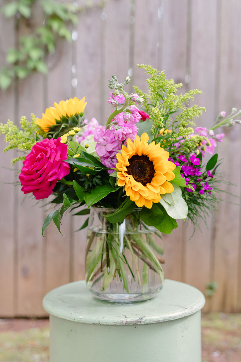 Vibrant mixed flower arrangement featuring yellow sunflowers, hot pink roses, light pink peonies, and purple blooms with green foliage in a clear glass vase on a sage pedestal