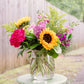 Vibrant floral arrangement with yellow sunflowers, fuchsia roses, pink spray flowers, and magenta blooms with chartreuse filler in a clear glass vase