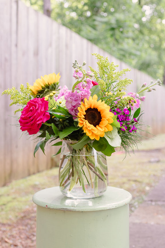 Vibrant floral arrangement with yellow sunflowers, fuchsia roses, pink spray flowers, and magenta blooms with chartreuse filler in a clear glass vase