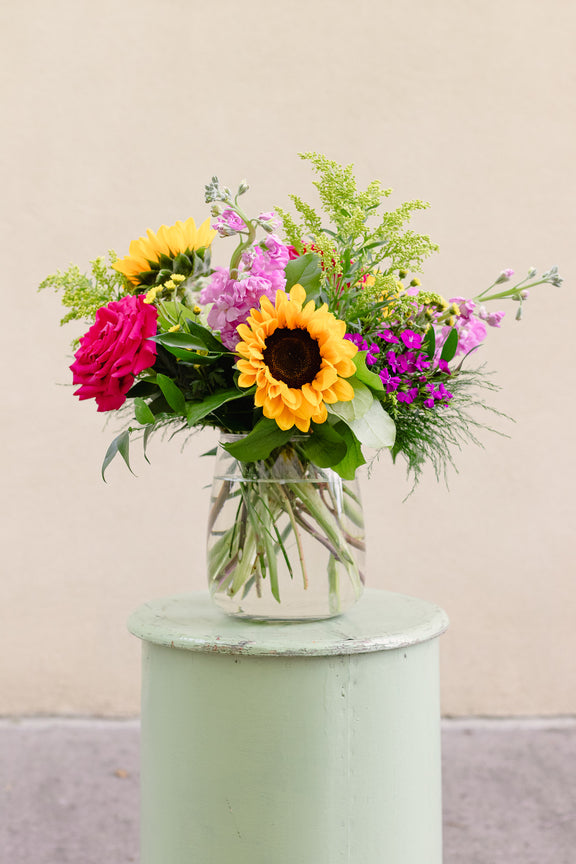 Colorful bouquet of flowers in a clear vase on a light green pedestal against a beige background