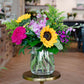 Vibrant floral arrangement in clear glass vase with yellow sunflowers, hot pink roses, pink carnations, and mixed greenery foliage.