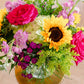 Vibrant floral arrangement with red roses, yellow sunflowers, pink and purple blooms, and green foliage in clear glass vase on wooden stand