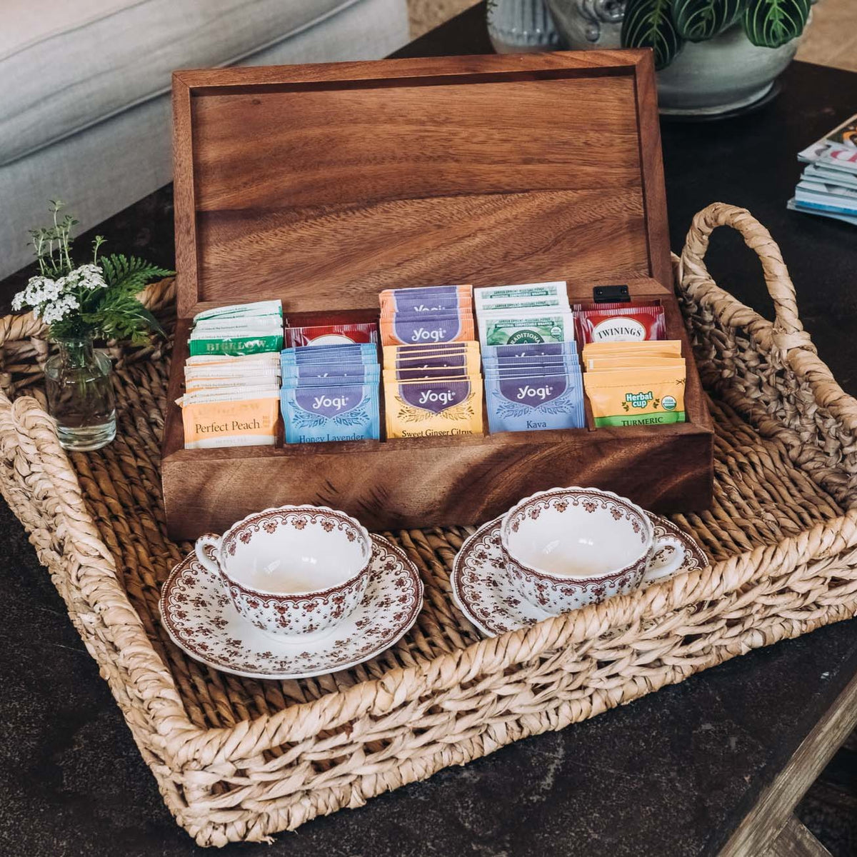 Open acacia wood tea box with 10 compartments displaying assorted tea boxes, set in woven basket with two patterned teacups and saucers