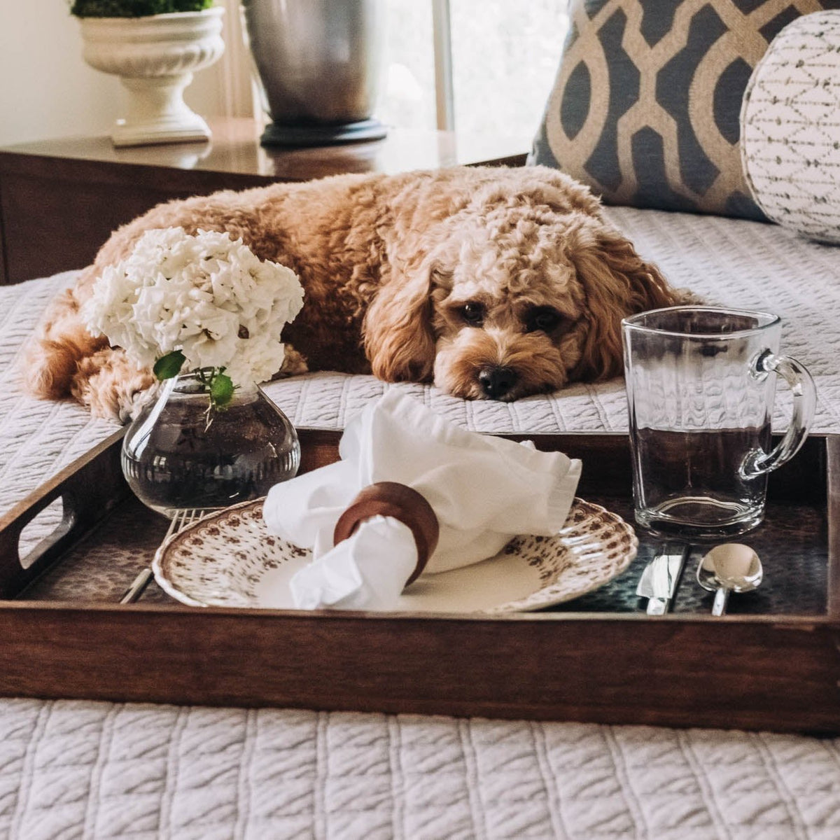 Rectangular brown wooden tray with hammered finish and inset handles, styled with white flowers, glassware, and cream napkin.