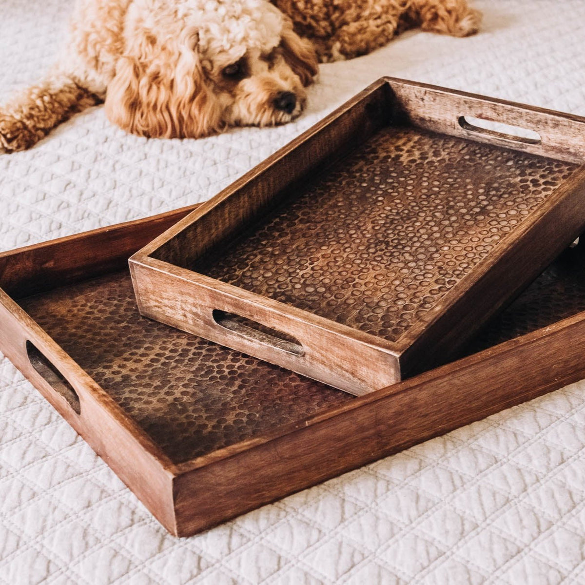 Two stacked rectangular wood serving trays with hammered bronze metallic inserts and side handles, displayed on white quilted surface