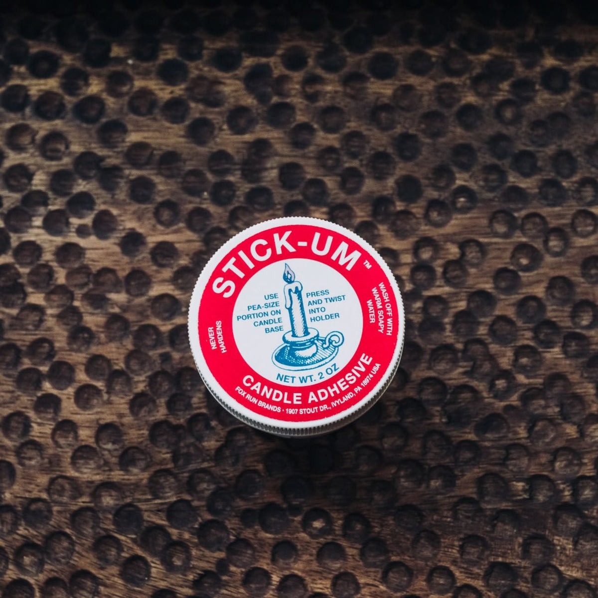 Red and white Stick-Um Candle Adhesive tin on leopard-print surface, product label showing 2 oz net weight with blue candle icon.
